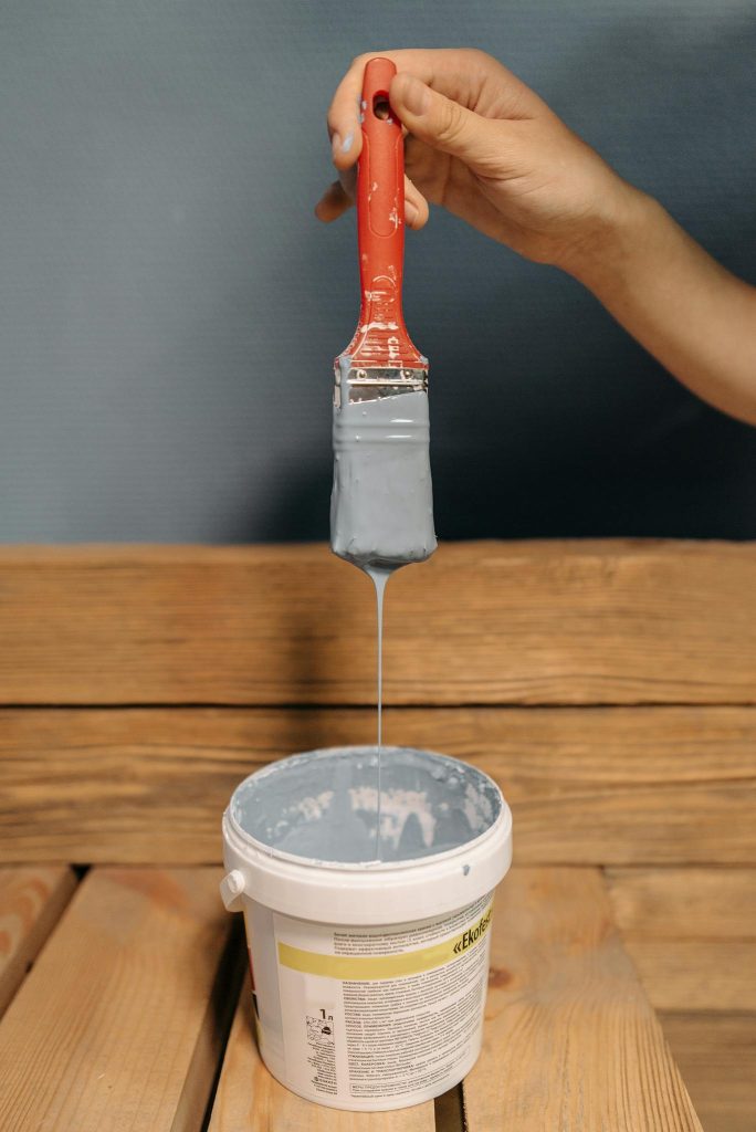 Close-up of a hand holding a paintbrush with gray paint dripping into a bucket on wooden surface.