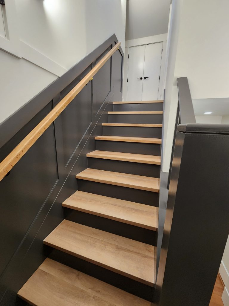 Modern interior staircase with dark painted paneling and railings, natural wood handrail and stair treads, and crisp white walls leading to double doors above.