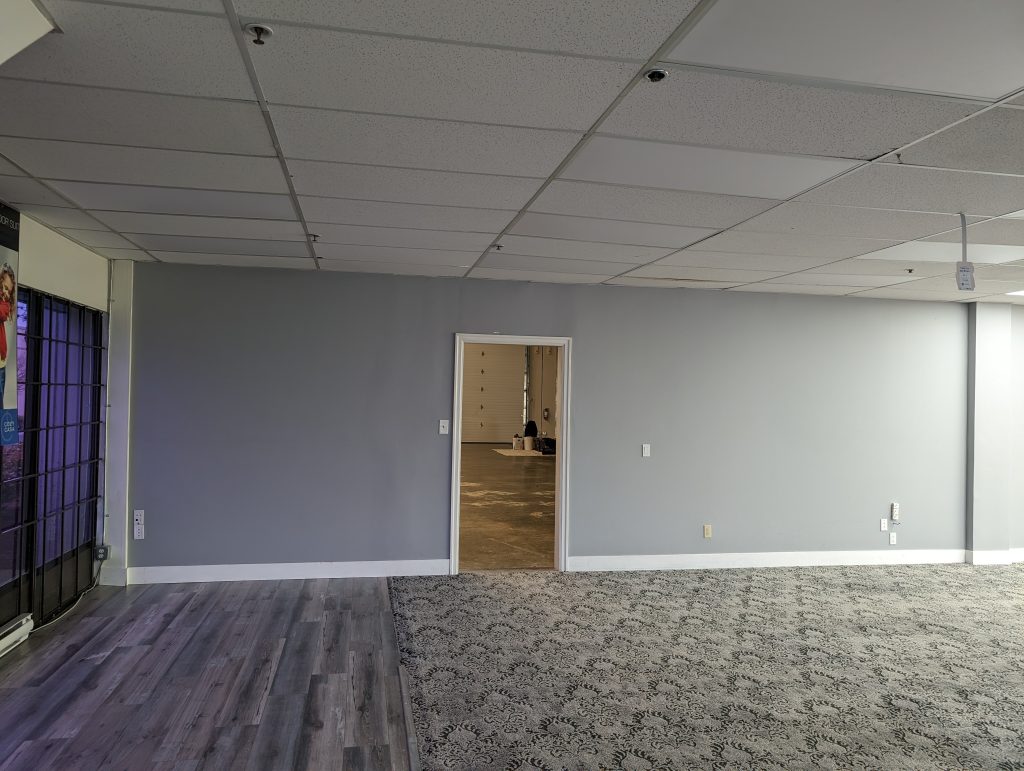 Open commercial interior with freshly painted soft grey walls, white trim, drop ceiling tiles, mixed carpet and vinyl flooring, and a doorway leading into a back workspace.
