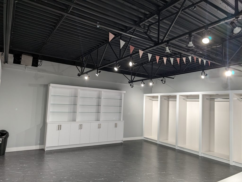 Freshly painted retail interior with light gray walls, white built-in display shelving, open merchandise bays, black exposed ceiling, and track lighting.