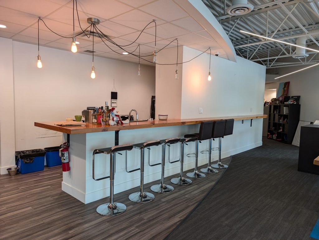 Modern commercial interior with freshly painted white walls, a long wood bar counter, pendant lighting, and bar stools in an open office lounge space.